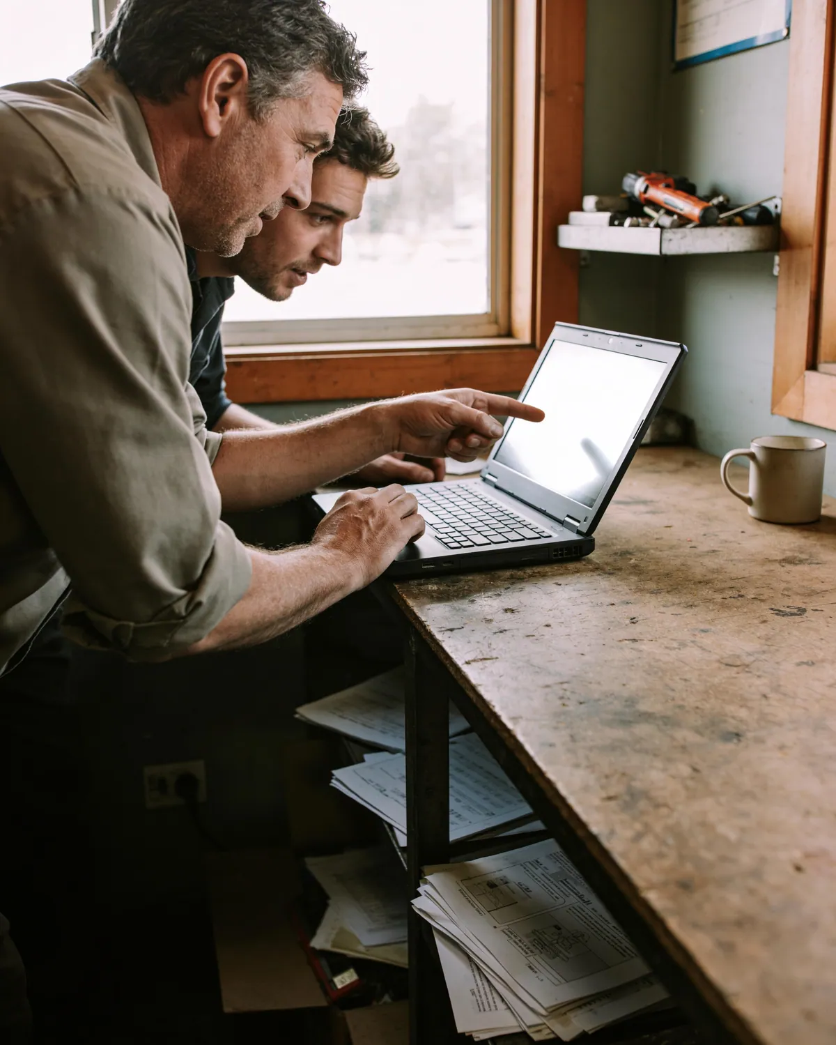 A business owner and a colleague leaning over a laptop on a worn wooden counter by a bright window, reviewing something together